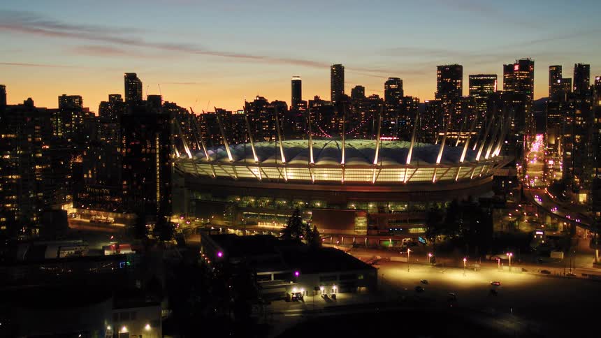 Aerial footage slowly pulls back from the brightly illuminated BC Place stadium against the stunning Vancouver city skyline at dusk