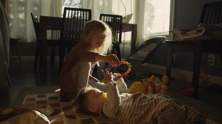 A toddler girl dangles a colorful rattle for her baby sibling as they play together on the floor in a sunlit room