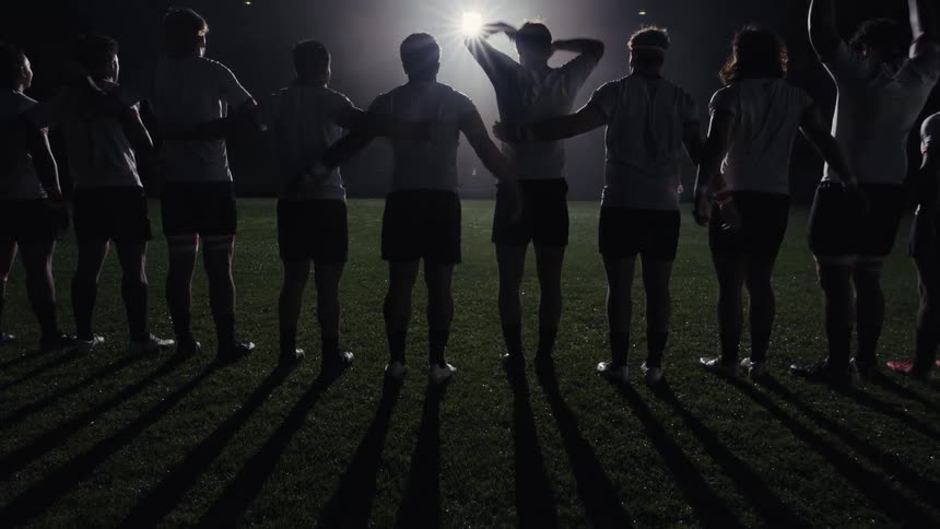 From behind, a rugby team unites with their arms around each other's shoulders, silhouetted by a single bright stadium light at night