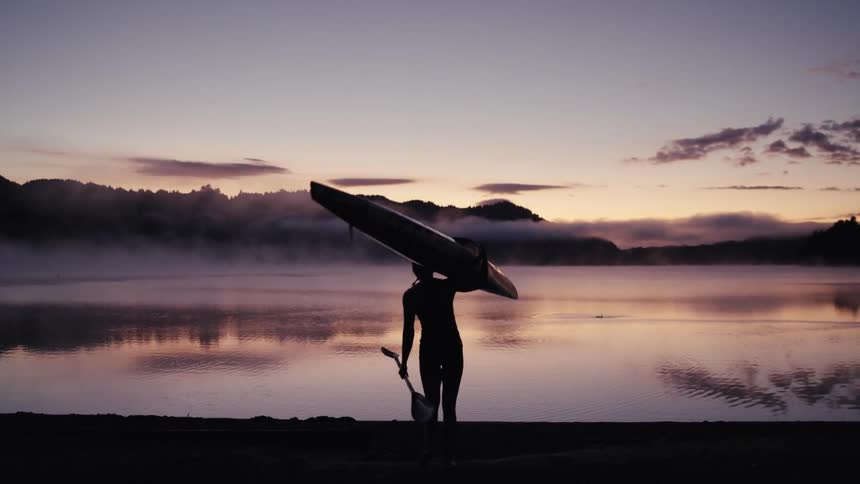 A woman in silhouette carries her kayak and paddle towards a serene and misty lake at sunrise