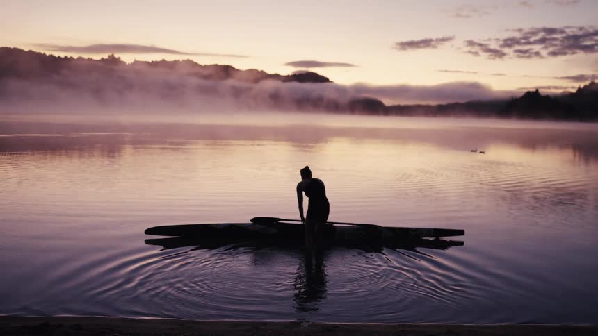 A silhouetted woman carries her kayak into a serene, misty lake at dawn and prepares for a peaceful morning paddle