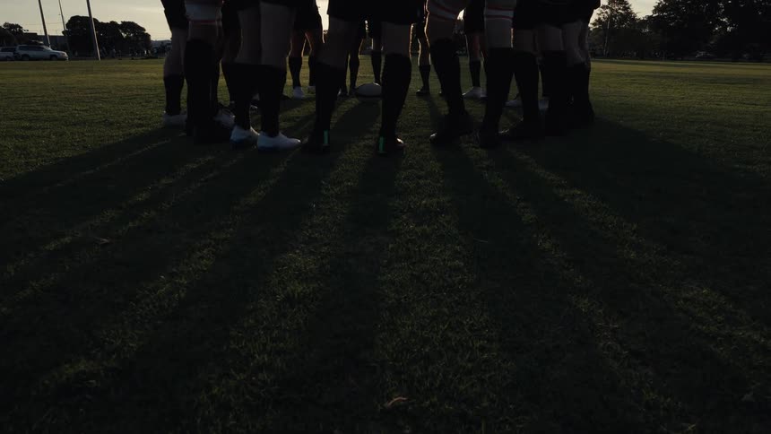 A sports team huddles together on a grassy field as the setting sun creates a dramatic starburst lens flare between their legs