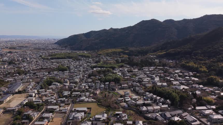 Aerial drone footage slowly pulls back over a quiet Japanese suburban town nestled at the base of lush green mountains