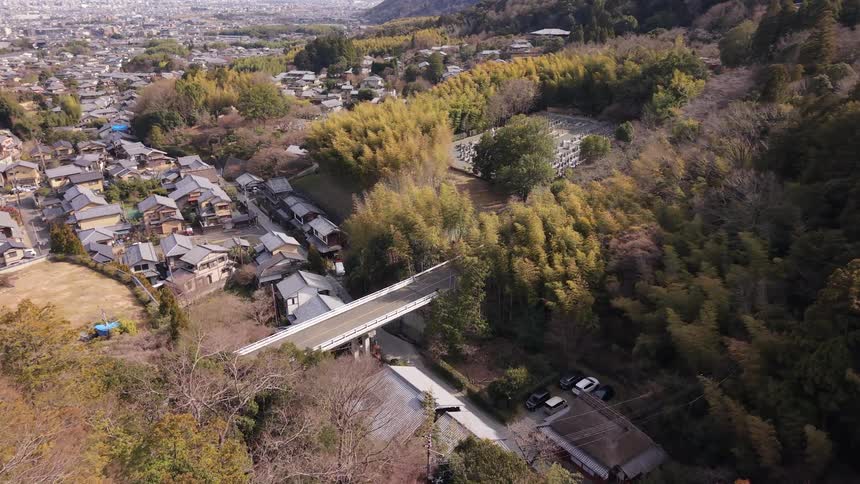 Aerial view flying over a forested valley towards a traditional Japanese village, with a modern road bridge and hillside cemetery