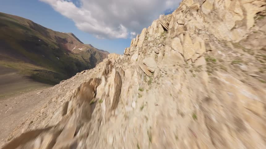 An exhilarating FPV drone flight soars up a rugged, rocky mountain face, skimming the ridgeline to reveal a majestic valley below