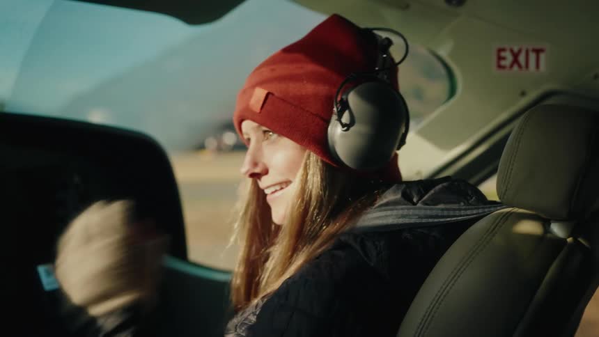 A smiling woman in a red beanie puts on her aviation headset in the cockpit of a small plane, ready for an adventure