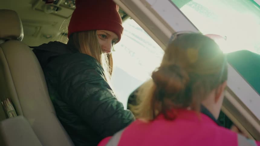 An excited female passenger wearing a red beanie sits in the cockpit of a small airplane and talks with her pilot before the flight