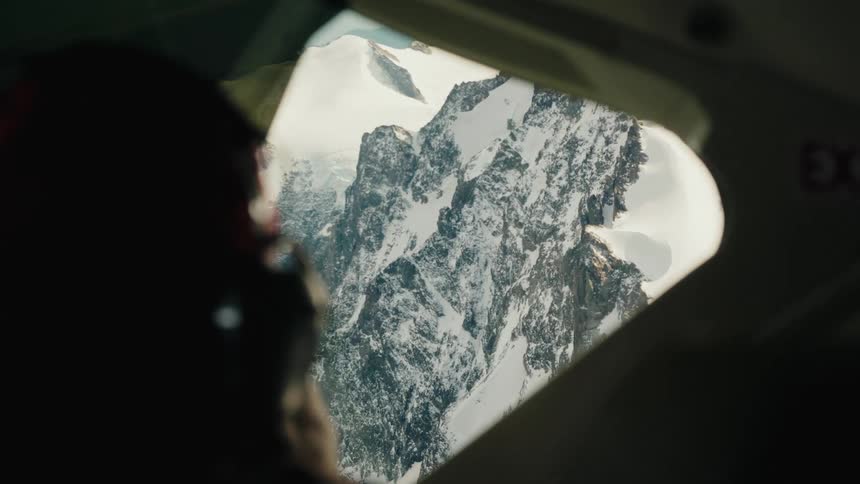 Over the shoulder shot of a person looking out of an airplane window at the majestic snow-capped mountains below