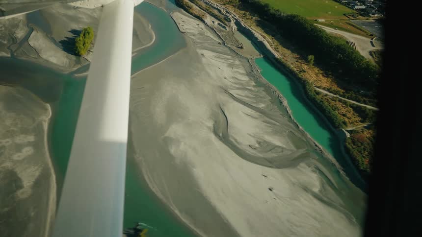 An aerial view from an airplane window shows a stunning turquoise braided river flowing through a wide, silty valley