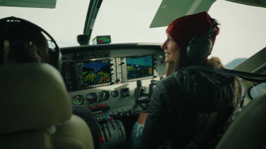 Two smiling women pilot a small airplane through a scenic mountain range as seen from inside the cockpit