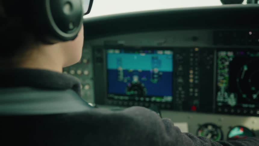 A pilot's hand adjusts a modern navigation system in a small aircraft cockpit before the camera pulls focus to her back