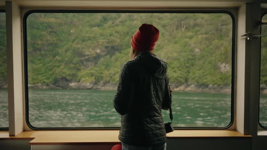 A woman in a red beanie stands inside a moving ferry, looking out the window at the passing fjord landscape