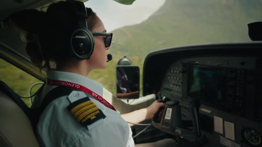 Over-the-shoulder view of a female pilot in uniform and sunglasses flying a small plane over a green mountainous landscape