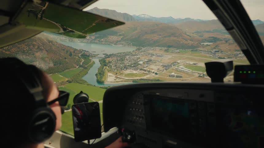 A female pilot in a cockpit flies a small aircraft, revealing a breathtaking view of a mountain town and lake below