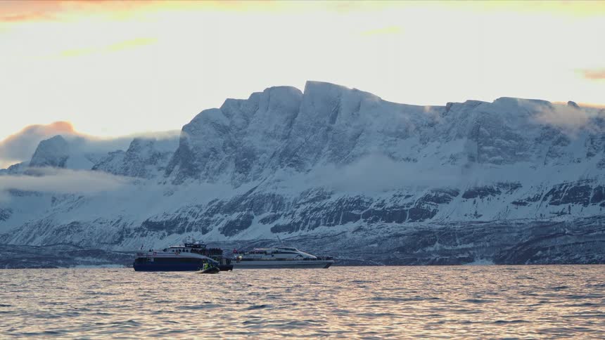 A serene wide shot of two boats floating in an arctic fjord with majestic snow-covered mountains in the background during sunset