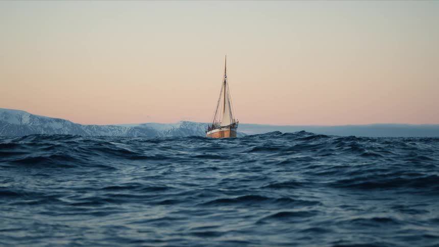 A classic sailboat sails on the vast, choppy ocean with snowy mountains in the distance during a beautiful sunset