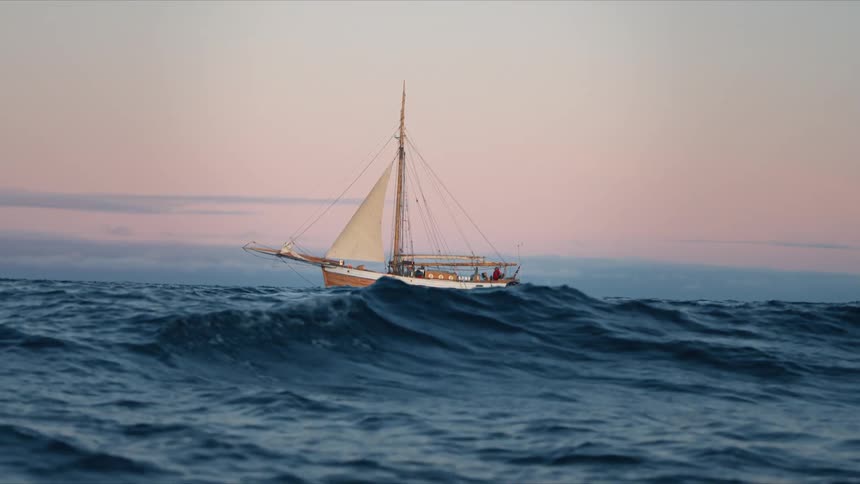 A classic wooden sailboat sails across rolling blue ocean waves under a serene pastel sunset sky