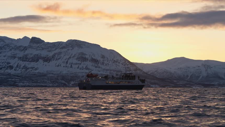 A tour boat filled with passengers floats on calm waters at sunset against a backdrop of majestic snow-covered mountains