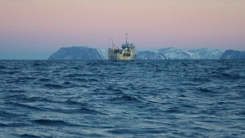 A fishing trawler floats on the vast, choppy blue ocean against a backdrop of snowy mountains and a pink sunset sky