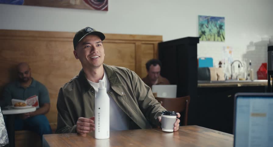 A friendly young Asian man joins someone at a table in a bustling coffee shop for a conversation