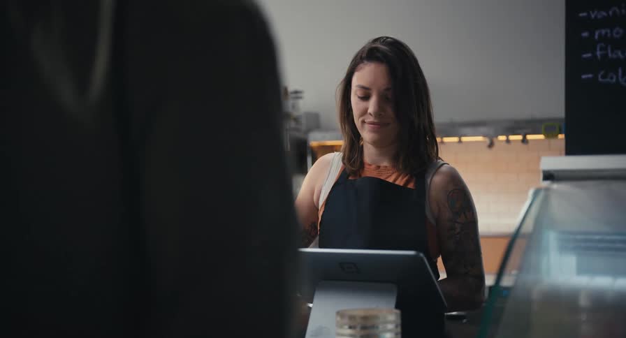A cheerful barista with tattoos smiles as she helps a customer at the counter of a cafe