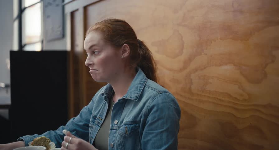 A young redhead woman works on her laptop and enjoys a snack while listening to music with wireless earbuds in a cozy cafe
