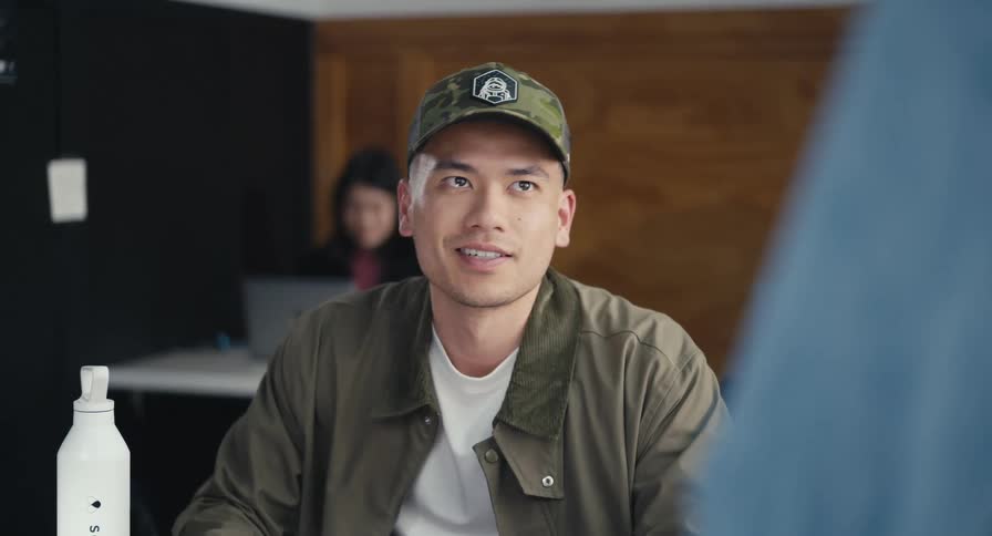 A young Asian man in a baseball cap attentively listens and contributes to a conversation in a collaborative workspace