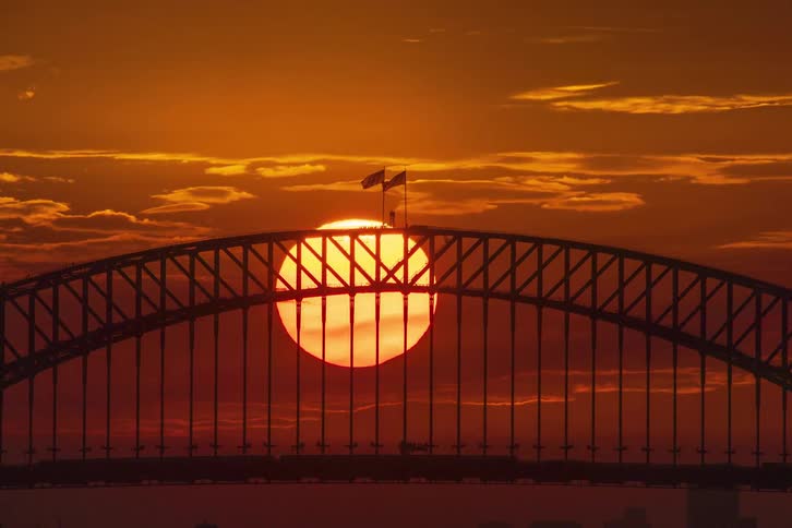 A dramatic timelapse shows the sun setting directly behind the silhouetted Sydney Harbour Bridge against a fiery orange sky