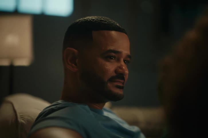 A man sits on a sofa in a dark room, listening attentively and smiling slightly