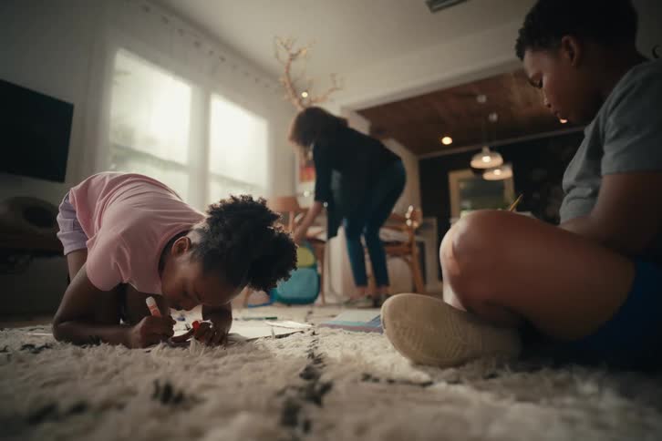 A young girl and her brother draw on the living room floor while their mother packs a backpack in the background