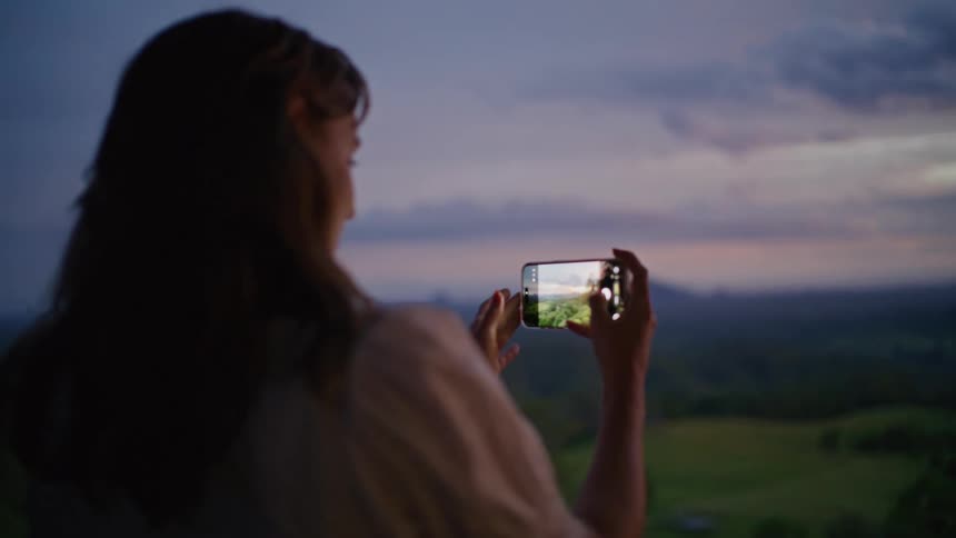 Over-the-shoulder shot of a woman taking a photo with her smartphone of the rolling hills at sunset