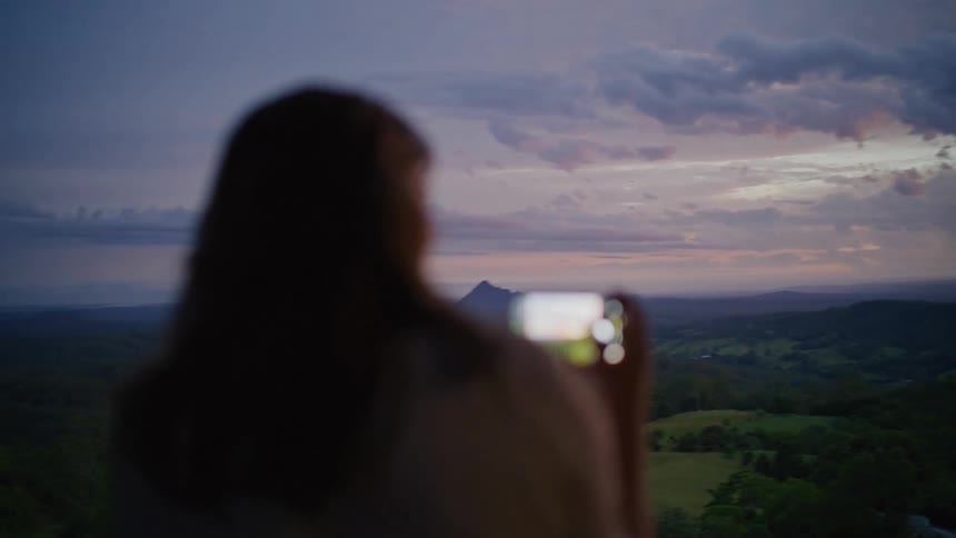 A woman captures a stunning mountain landscape on her smartphone during a colorful twilight, with the focus shifting to the phone's screen