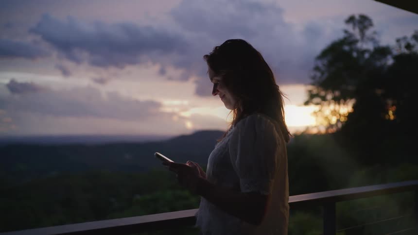 A young woman stands on a balcony and uses her smartphone as the sun sets over a mountain valley