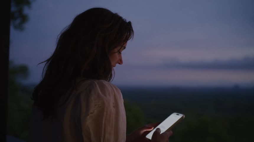 A woman smiles while using her phone at dusk, as the focus shifts to reveal a beautiful mountain landscape in the background