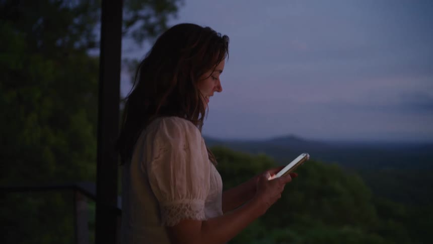 A smiling woman scrolls on her smartphone on a serene balcony at twilight, with a vast mountain landscape in the background