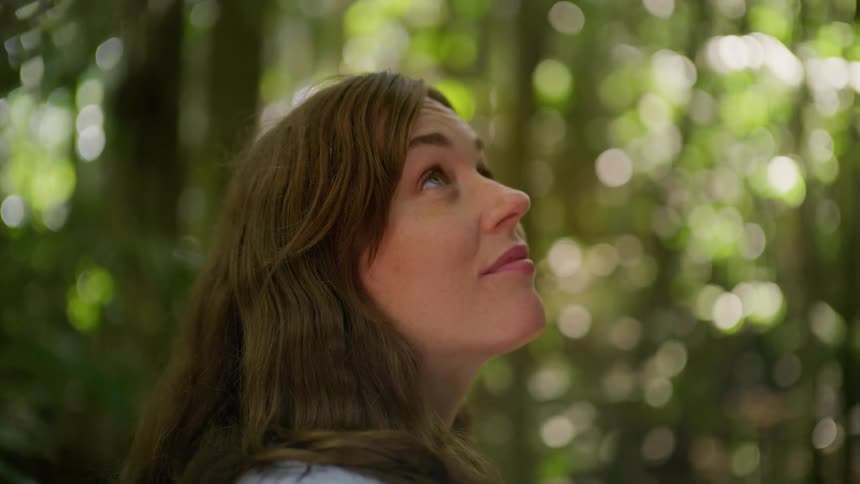 A young woman peacefully connects with nature, closing her eyes and smiling softly in a tranquil, sun-dappled forest