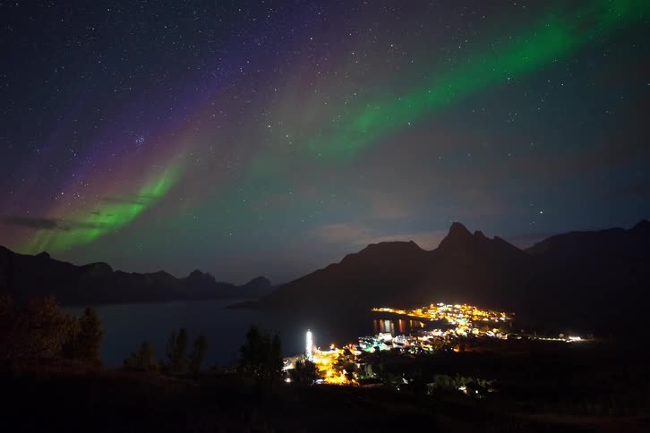 A stunning time-lapse captures the colorful Aurora Borealis dancing in the starry night sky over a tranquil coastal town nestled among mountains