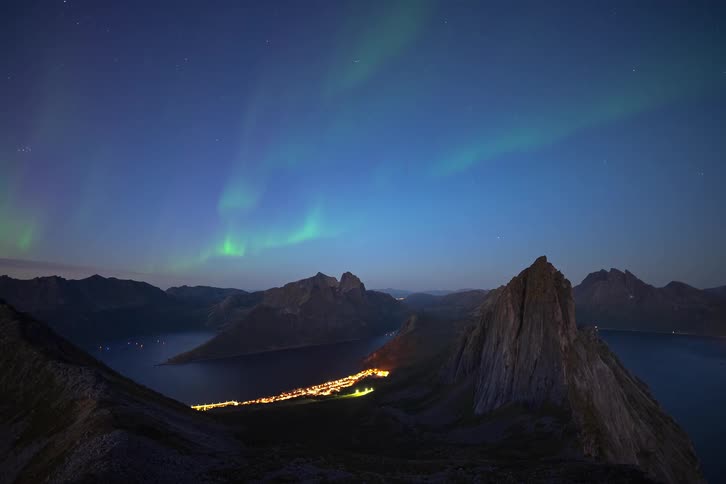 Majestic time-lapse of the Aurora Borealis glowing green above a coastal village nestled between towering mountains and a fjord at night