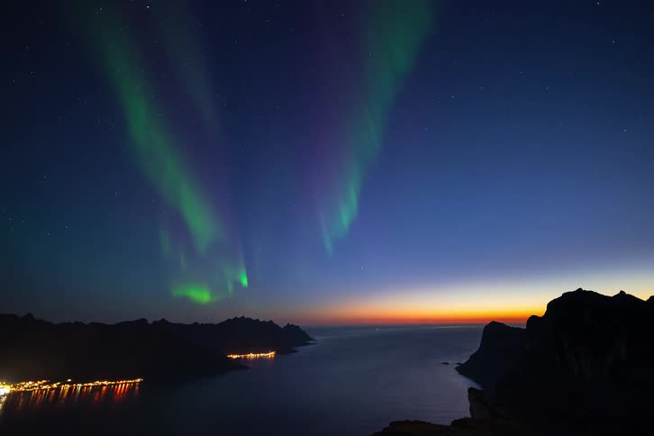 A stunning time-lapse shows the vibrant green Aurora Borealis dancing in the starry night sky over a tranquil sea and a lit coastal town nestled among mountains