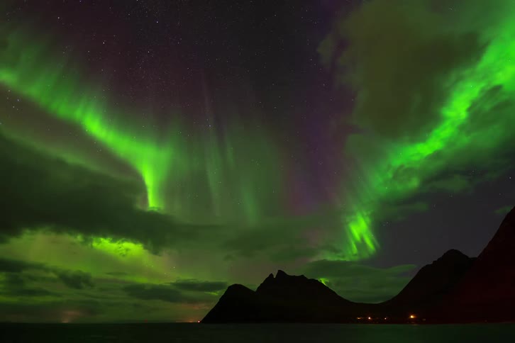 A mesmerizing time-lapse captures the vibrant green and purple hues of the Aurora Borealis dancing across the starry night sky above a silhouetted mountain and the ocean