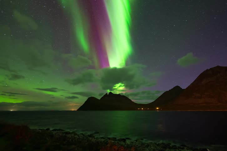 A stunning time-lapse shows the green and purple Aurora Borealis dancing in the starry night sky over a mountainous coastal landscape