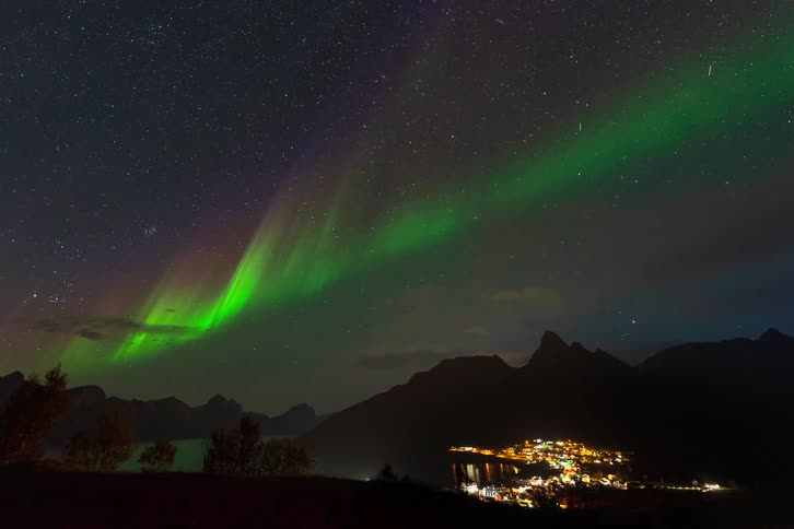 A mesmerizing time-lapse of the vibrant green and purple Aurora Borealis dancing in the starry night sky over a silhouetted mountain range and a glowing coastal town
