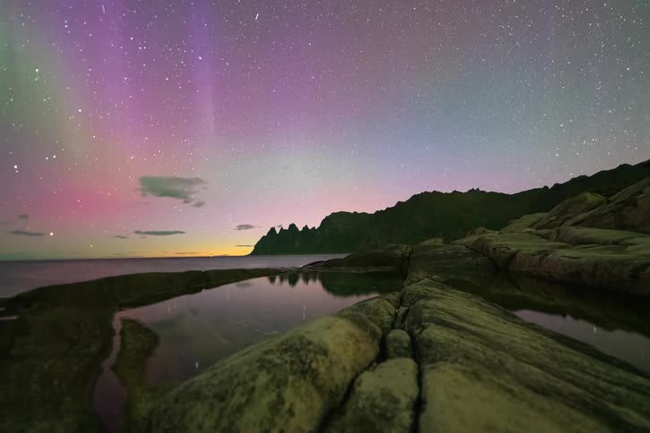 A vibrant timelapse captures the pink and green Aurora Borealis dancing across a star-filled sky above a rocky coastline