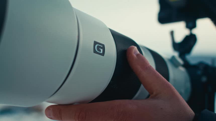A filmmaker's hand adjusts the focus ring on a large white telephoto lens mounted on a tripod outdoors