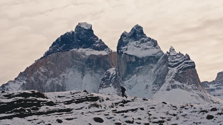 A lone hiker in slow motion traverses a snowy mountain landscape with the majestic twin peaks of a vast mountain range in the background