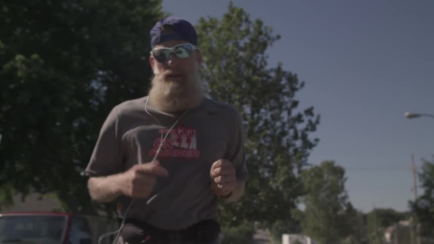 A man and a purple hat sunglasses and a gray shirt jogging on a sunny day