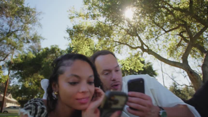 A young, stylish couple relaxes together on a blanket in a sunny park, scrolling on their smartphones
