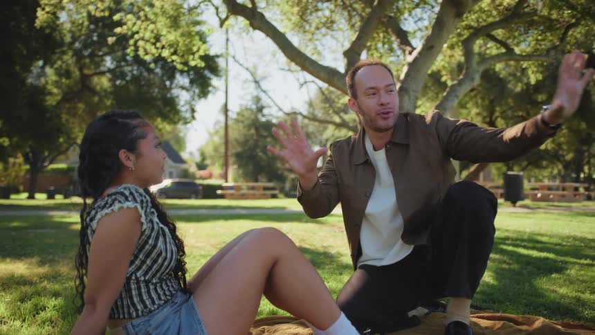A young man animatedly tells a story to a woman as they enjoy a picnic in the park on a sunny day