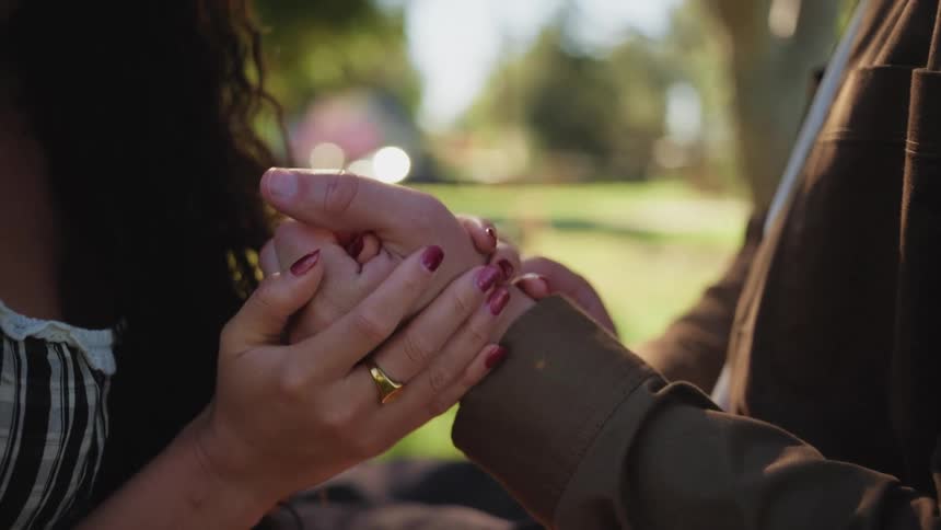 An intimate close-up shot of a woman with red nails affectionately holding her lover's hand in a sunny park