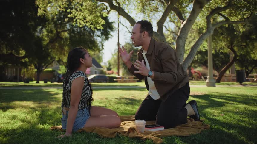 A man animatedly tells a story to his girlfriend as they enjoy a sunny day having a picnic in the park